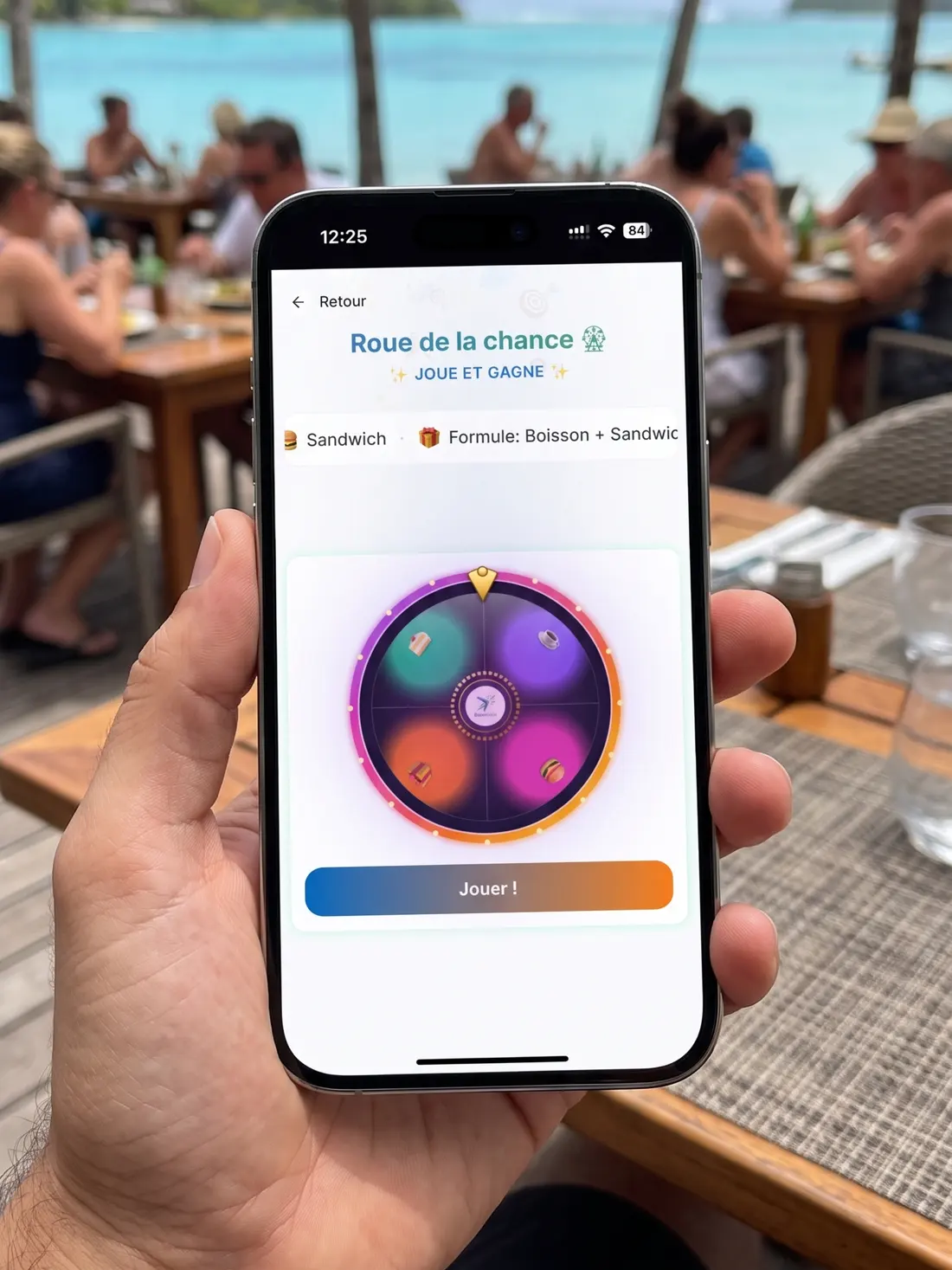 Un client tient son téléphone à table, face au lagon : la webapp BoumRank affiche la roue de la chance et le bouton « Jouer ! ».