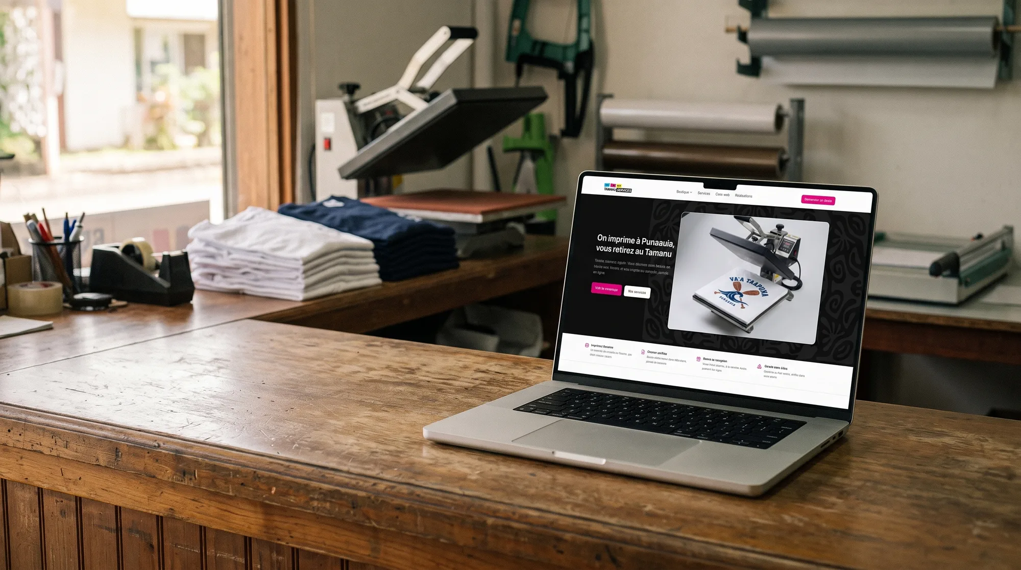 La page d'accueil de Tamanu Services ouverte sur un ordinateur portable, posé sur le comptoir en bois de l'atelier, presse à chaud et t-shirts vierges derrière.